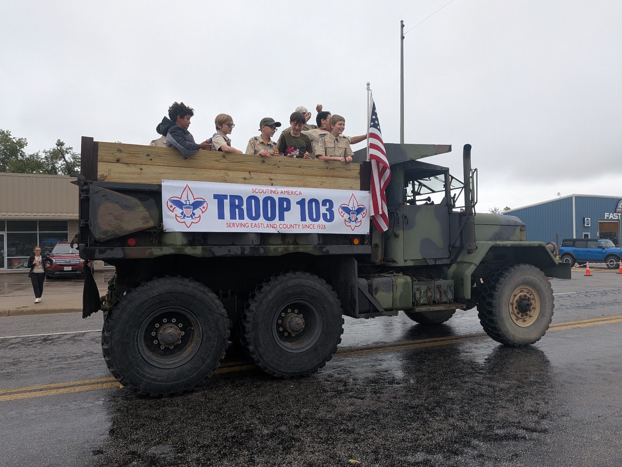 Troop 103 participants in a camouflage parade truck displaying the troop banner and an American flag.